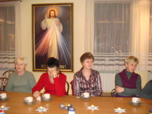 Grupa osób siedzi przy stole w pomieszczeniu, za nimi wisi obraz religijny.