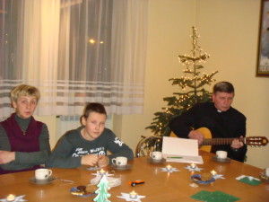 Trzy osoby siedzą przy świątecznym stole, mężczyzna gra na gitarze, w tle widoczna choinka i okno.