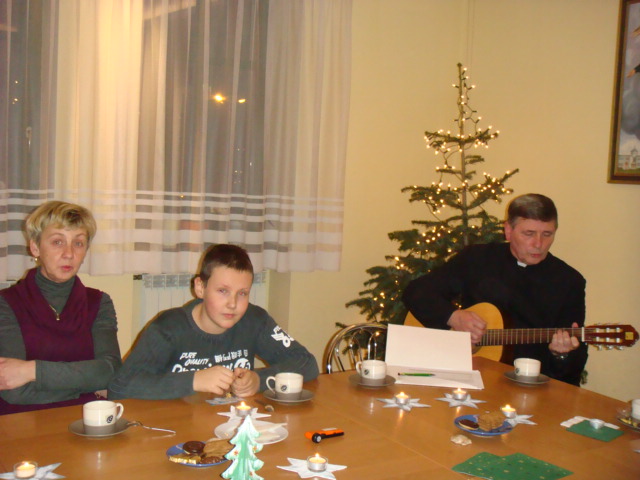Trzy osoby siedzą przy świątecznym stole, mężczyzna gra na gitarze, w tle widoczna choinka i okno.
