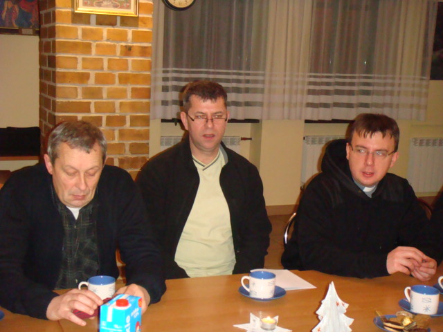 Trzej mężczyźni siedzą przy stole, na którym stoją filiżanki i serwetki, w pomieszczeniu z ceglanym kominkiem.