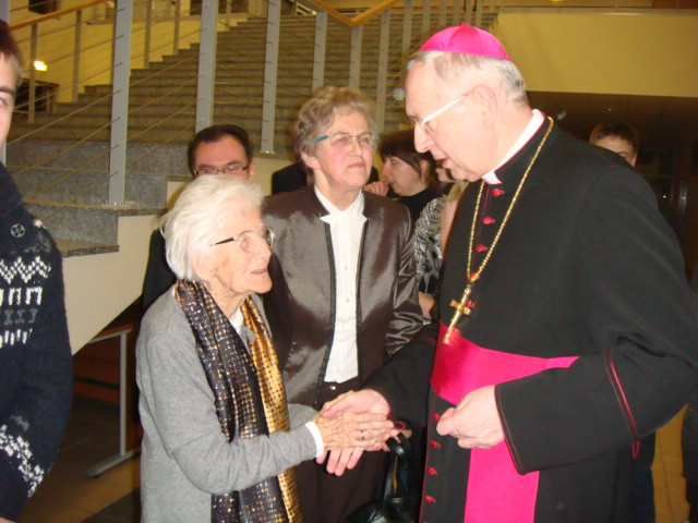 Biskup w stroju liturgicznym wita kobiety podczas wydarzenia w galerii, w tle widoczne schody.