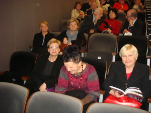 Grupa osób siedzi w widowni teatru lub sali koncertowej, w rzędach krzeseł.