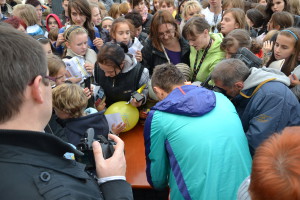 Osoba w kurtce turkusowej podpisuje piłkę, otoczona tłumem osób podczas wydarzenia w 2012 roku.
