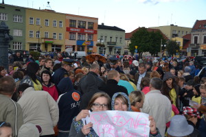 Tłum ludzi zgromadzony na placu miejskim, w tle budynki z witrynami sklepów i drzewa.