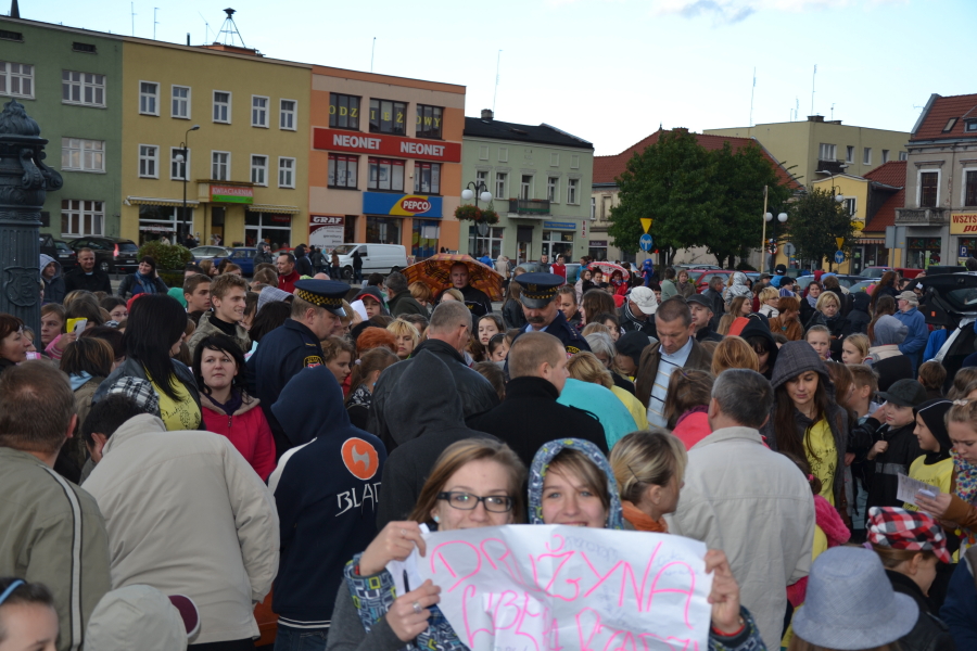 Tłum ludzi zgromadzony na placu miejskim, w tle budynki z witrynami sklepów i drzewa.