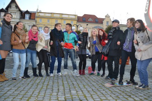 Grupa osób stoi na brukowanym placu, niektórzy na deskorolkach, w tle widoczne budynki.
