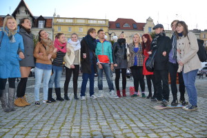 Grupa osób stoi na brukowanym placu przed budynkami.