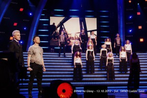 Scena koncertowa z wykonawcami i jury, oświetlona niebieskimi światłami, z napisem „Bitwa na Głosy” z datą 13.10.2012.
