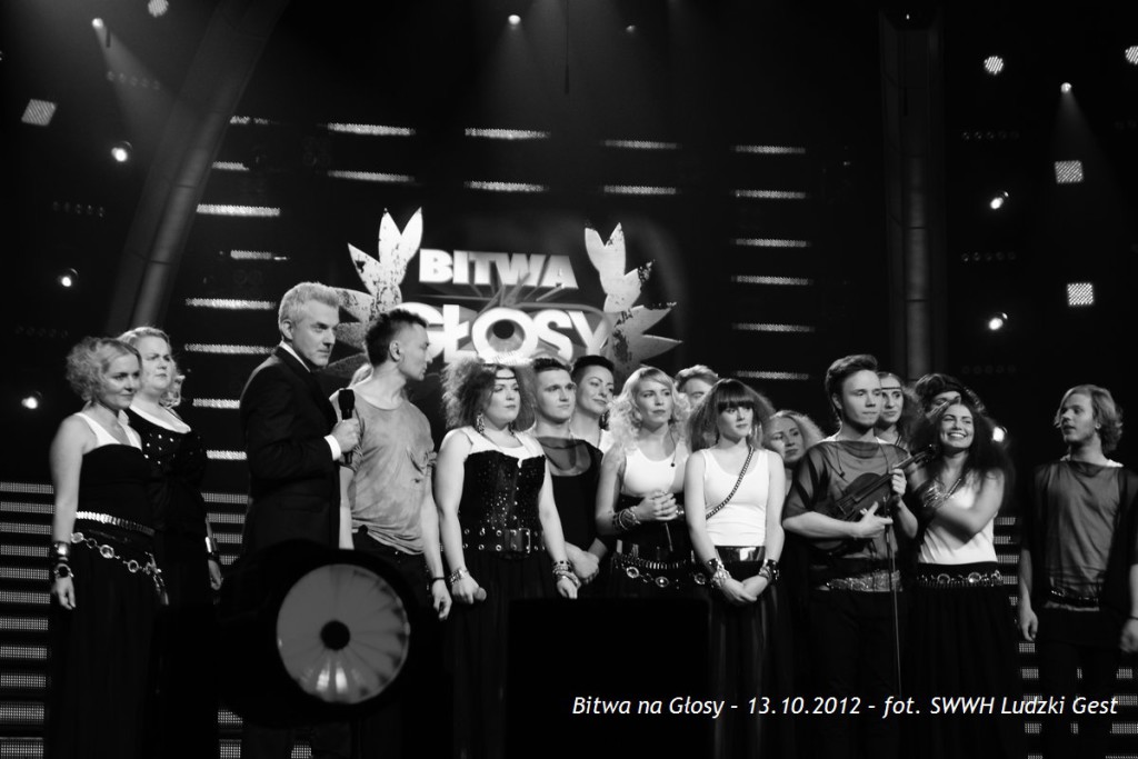 Grupa osób stoi na scenie, oświetlona reflektorami, przed napisem „Bitwa na Głosy”.