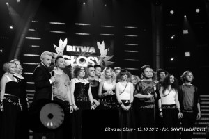 Grupa osób na scenie, oświetlona reflektorami, z widocznym logotypem „Bitwa na Głosy” w tle.