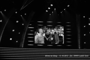 Dwóch mężczyzn siedzi na scenie przed publicznością, w tle widoczne oświetlenie i elementy dekoracji scenografii.