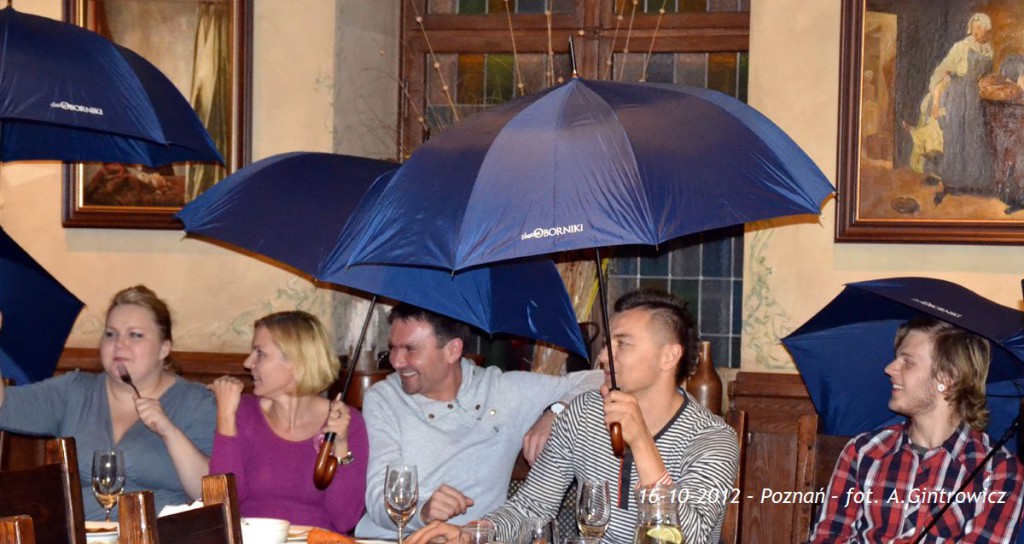 Grupa osób siedzi przy stole w restauracji, trzymając niebieskie parasole.