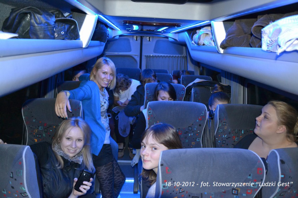 Grupa osób podróżuje autobusem z niebieskim oświetleniem, widoczne siedzenia i wnętrze pojazdu.