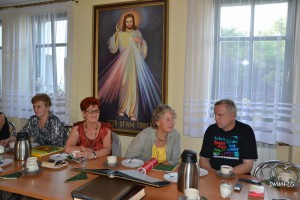 Grupa osób siedzi przy stole w pomieszczeniu, za nimi wisi obraz przedstawiający Jezusa Miłosiernego.