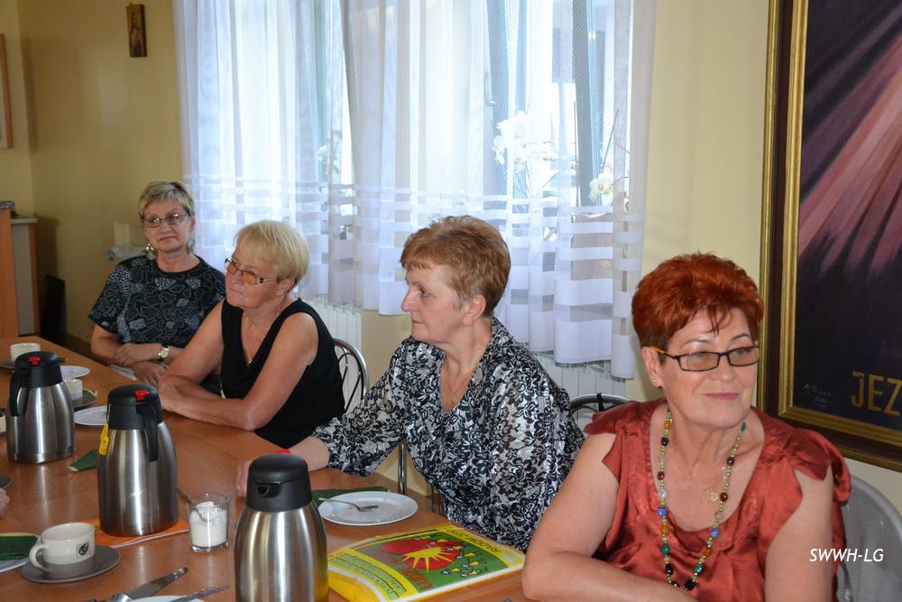 Cztery osoby siedzą przy stole w pomieszczeniu z dużymi oknami i widocznym obrazem na ścianie.