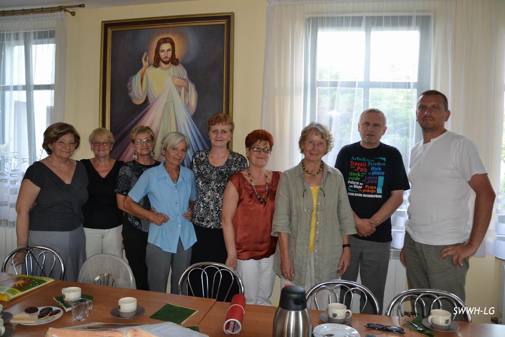 Grupa osób stoi przy stole w pomieszczeniu z dużym obrazem religijnym i oknem z zasłonami.