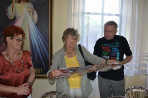 Osoby rozmawiają w pomieszczeniu z dekoracją religijną i plakatem wielojęzycznym.