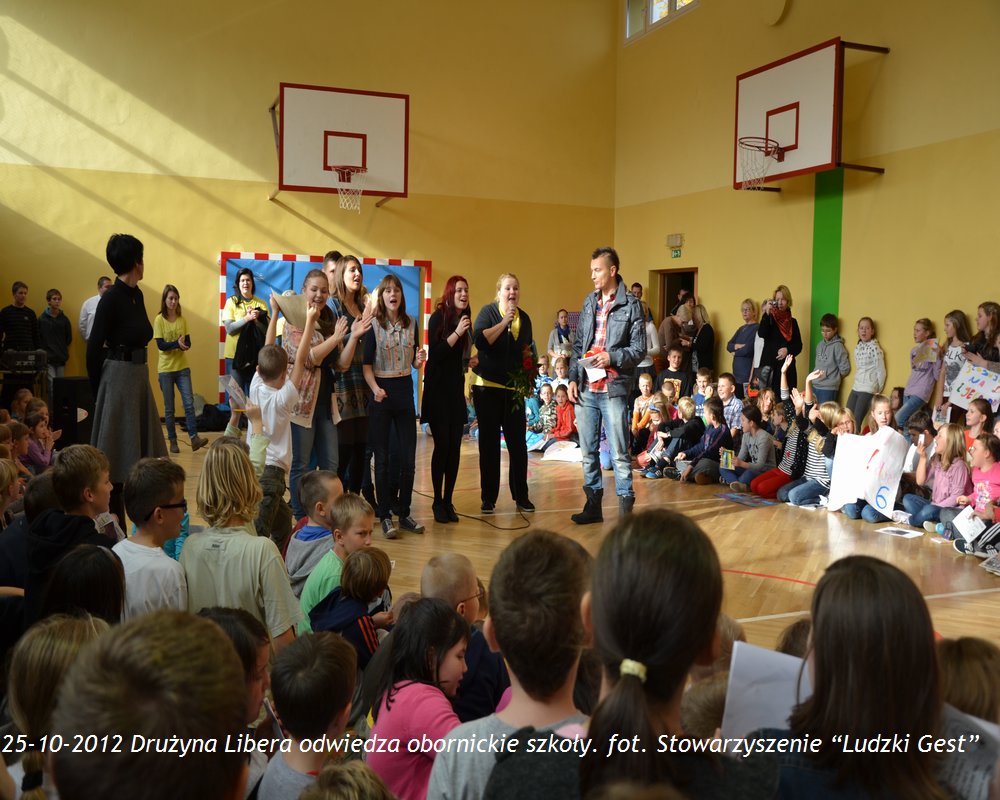 Grupa osób występuje przed publicznością dzieci w szkolnej sali gimnastycznej, 25 października 2012.