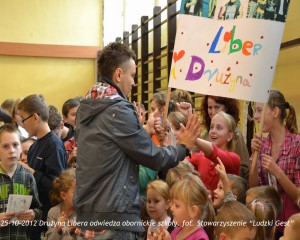 Mężczyzna w kurtce wita dzieci w szkole, trzymając ręce wysoko. Na plakacie widnieje napis „Liber Drużyna”.