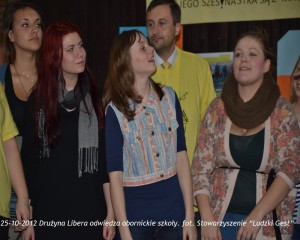 Grupa osób uczestniczy w spotkaniu w pomieszczeniu, w tle widoczne tablice informacyjne.