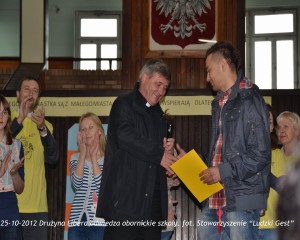 Grupa osób uczestniczy w spotkaniu w obornickiej szkole, w tle widoczny Polski orzeł i tablica informacyjna.