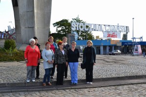 Grupa osób stoi na chodniku przed budynkiem z napisem „Stocznia Czysta”, w tle widoczna konstrukcja betonowa.