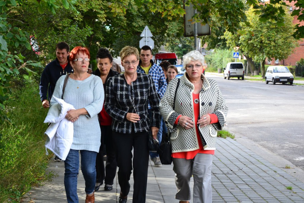 Grupa osób idzie chodnikiem wzdłuż drogi, w tle widoczne drzewa i budynki.