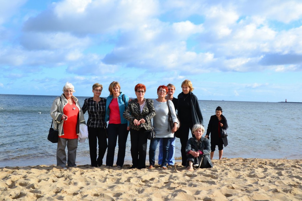 Grupa osób stoi na piaszczystej plaży przy morzu w słoneczny dzień.