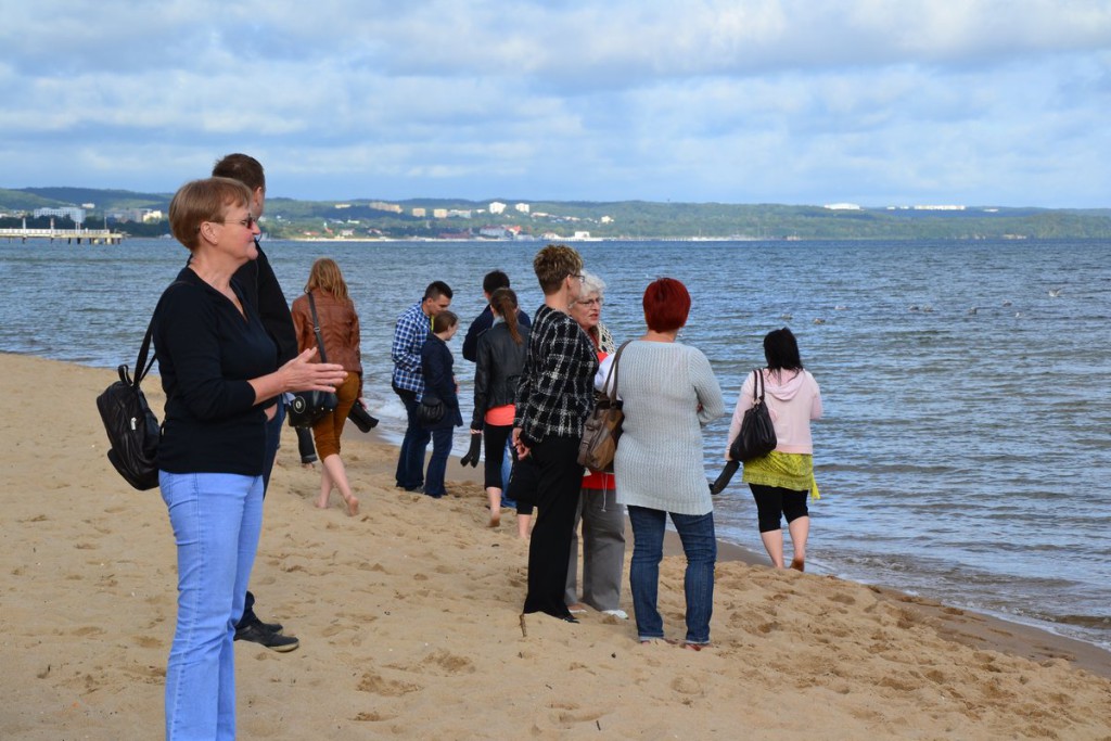 Grupa osób uczestniczy w spotkaniu na piaszczystej plaży, w tle widoczna woda i linia brzegowa.