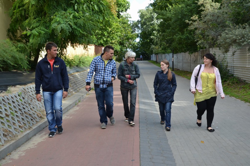 Pięć osób idzie po brukowanym chodniku, otoczone zielenią drzew i krzewów, w tle widoczne budynki.
