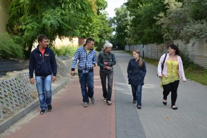 Pięć osób idzie po brukowanym chodniku, otoczone zielenią drzew i krzewów, w tle widoczne budynki.