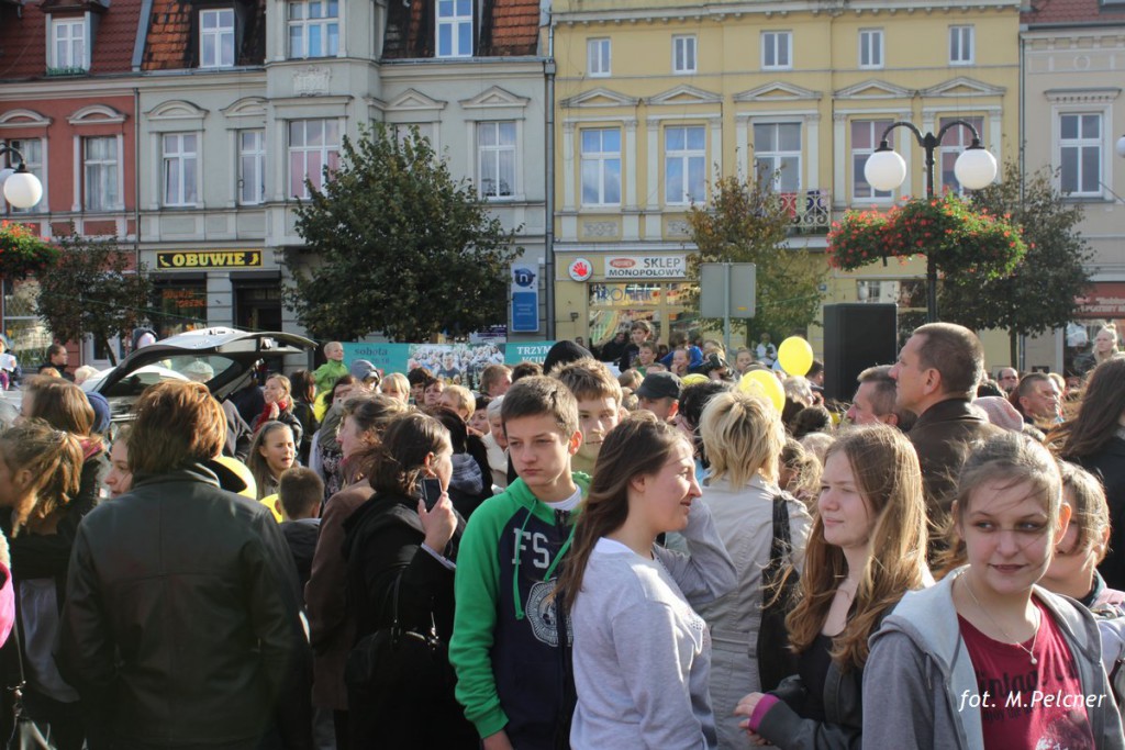 Tłum ludzi przemieszcza się po placu miejskim, w tle widoczne budynki z witrynami i drzewa.