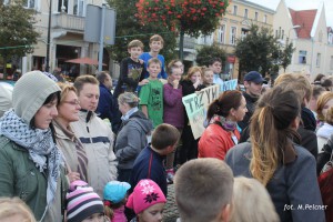 Tłum ludzi uczestniczy w wydarzeniu na placu miejskim, w tle budynki i drzewa.