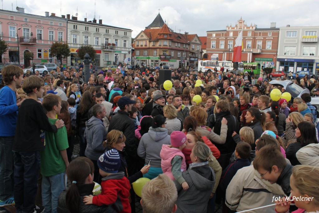 Tłum ludzi zgromadzony na placu miejskim, w tle budynki i drzewa, w rękach balony.