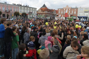 Tłum ludzi zgromadzony na placu miejskim, w tle budynki i drzewa, w rękach balony.