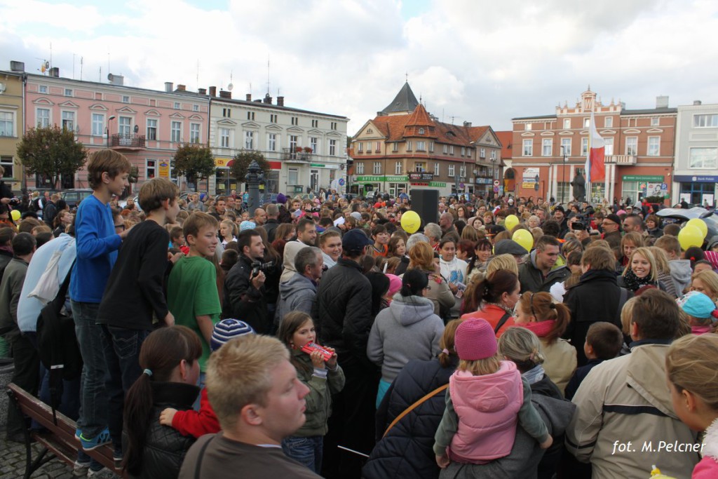 Tłum ludzi zgromadzony na placu miejskim, w tle budynki i niebo z chmurami.