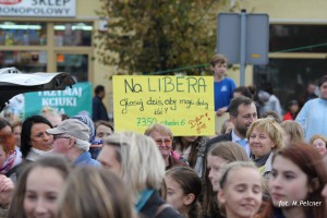 Uczestnicy wydarzenia trzymają transparent z napisem „Na LIBERA” i liczbami, w tle widoczny sklep.