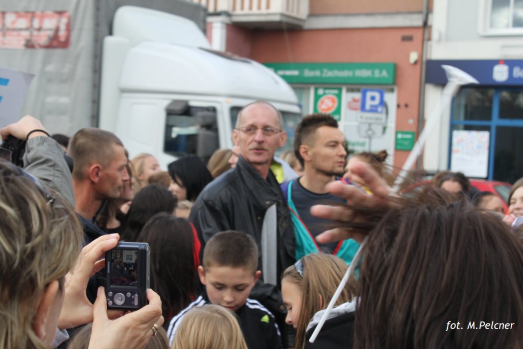 Tłum ludzi robi zdjęcia osobie w otoczeniu ochroniarzy, na tle budynków i pojazdu.