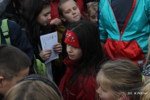 Grupa dzieci w kurtkach uczestniczy w wydarzeniu plenerowym, trzymając w rękach dokumenty.