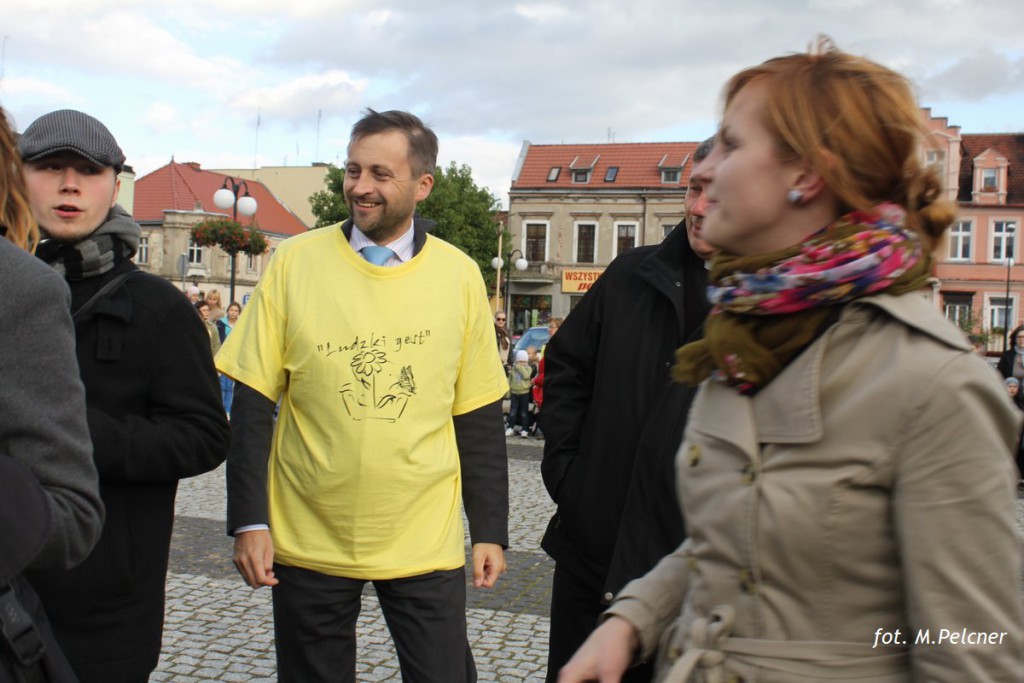 Mężczyzna w żółtej koszulce stoi w tłumie na placu, w tle widoczne budynki i lampy.