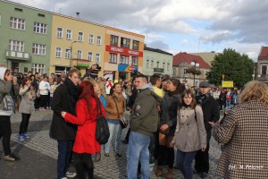 Tłum ludzi przemieszcza się po brukowanym placu miejskim, w tle widoczne budynki z witrynami sklepów.
