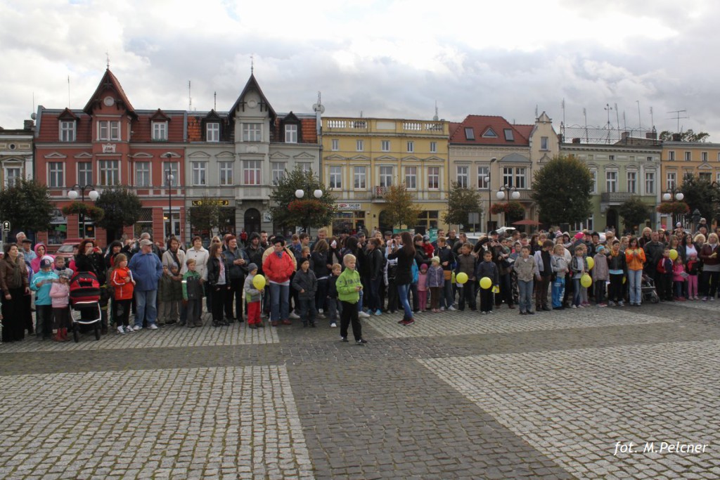 Grupa osób uczestniczy w wydarzeniu na placu, otoczonym budynkami z czerwonymi dachami.