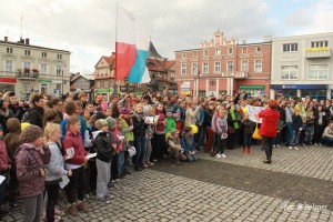 Grupa dzieci i dorosłych zgromadzona na placu brukowanym, z flagą i pomnikiem w tle.