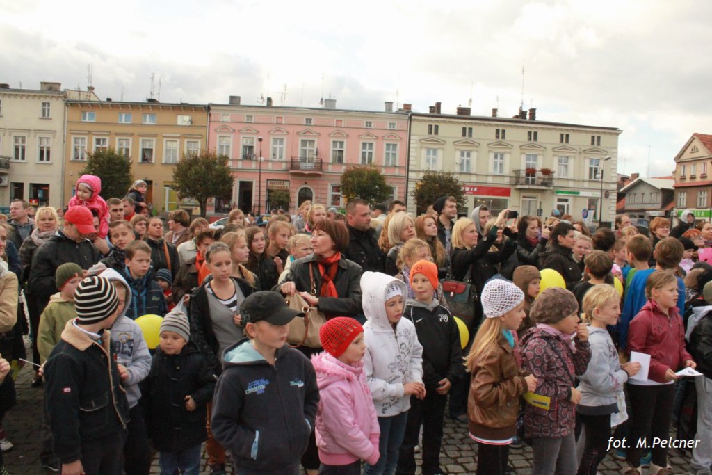 Tłum ludzi zgromadzony na placu, w tle budynki i drzewa.
