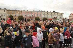 Tłum ludzi zgromadzony na placu, w tle budynki i drzewa.
