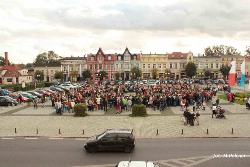 Tłum ludzi zgromadzony na placu przed zabytkowymi kamienicami, w tle widoczne drzewa i pochmurne niebo.
