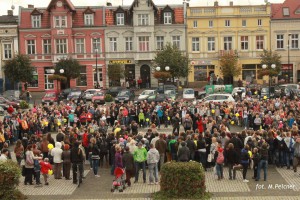 Tłum ludzi zgromadzony na placu, otoczony budynkami z czerwonymi dachami i witrynami sklepów.