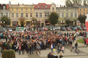 Tłum ludzi zgromadzony na placu miejskim, w tle budynki z witrynami i polska flaga.