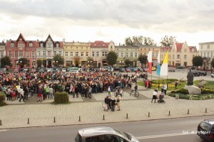 Tłum ludzi zgromadzony na placu miejskim, otoczonym zabytkowymi kamienicami i drzewami.