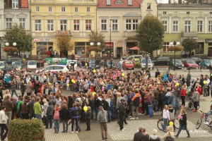 Tłum ludzi zgromadzony na placu, otoczony budynkami z witrynami sklepów i samochodami.